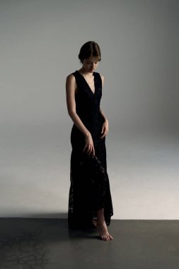 Siyah gece elbisesi içindeki zarif kadın dramatik ışıklandırmalı minimalist stüdyo arkaplanına karşı zarifçe poz veriyor.