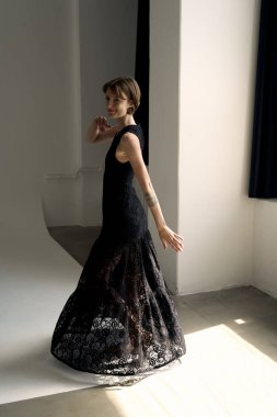 Dantelli siyah elbiseli model modern stüdyoda doğal pencere ışıklandırmasıyla zarifçe poz veriyor.