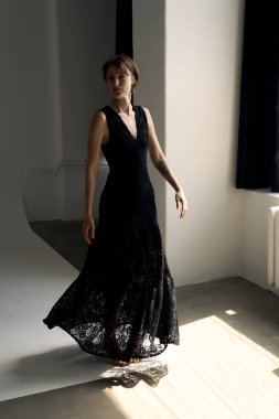 Akan siyah gece elbisesi içindeki zarif kadın dramatik gölgelerle modern mimari stüdyo alanında zarifçe ilerler.