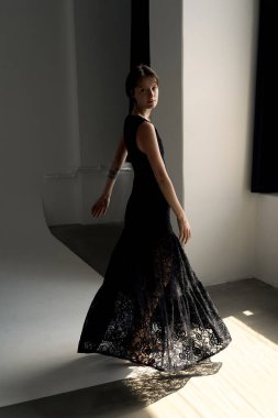 Zarif siyah gece elbisesi içindeki profesyonel model kıvrımlı mimari detaylarla parlak minimalist stüdyoda güven içinde poz veriyor.