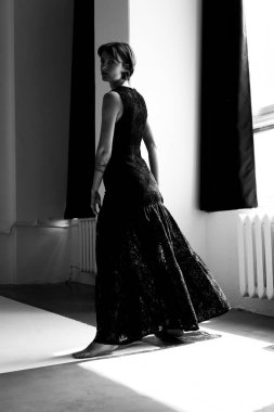 Dalgalı siyah elbiseler içindeki zarif kadın modern beyaz stüdyo alanında doğal ışıklandırmayla zarifçe hareket ediyor.