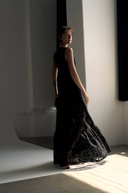Dalgalı siyah elbisesi içindeki zarif kadın doğal ışıklandırmayla dramatik beton mimari alanlarda zarif bir şekilde ilerler.
