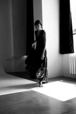 Moda modeli akıcı siyah elbise modern beton mimari alanda doğal pencere ışığı ile dramatik siluet oluşturur.