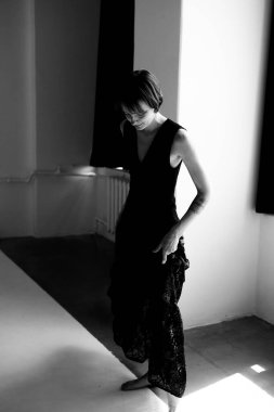 Dalgalı siyah resmi elbise içindeki zarif kadın modern beton stüdyo alanında doğal aydınlatmayla zarifçe hareket ediyor.