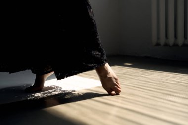 Zarif siyah elbiseli adam, parlak minimalist iç mekanda, güneş ışığıyla aydınlanmış ahşap zeminde yürüyor.