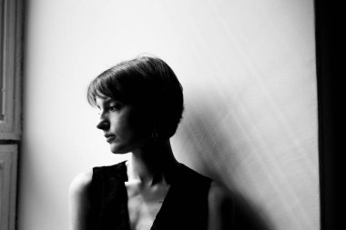 Siyah ceketli, kısa saçlı, beyaz duvarlı bir kadının dramatik silueti.