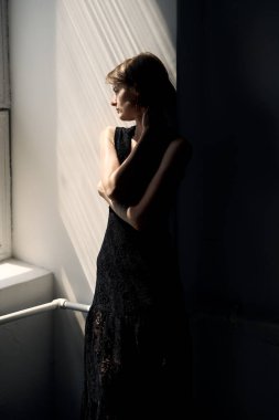 Siyah elbiseli kadın minimalist iç dekorda parlak pencerenin önünde duruyor.