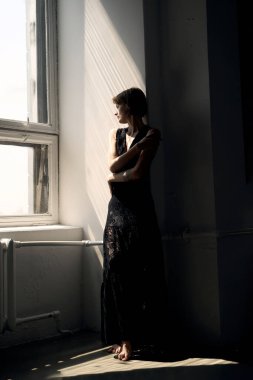 Siyah elbiseli bir kadın kapıda duruyor. Pencere ışığı içeri giriyor.