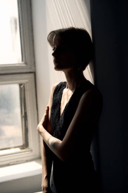 Siyah kolsuz bluzlu kadın minimalist iç mekandaki parlak pencerenin önünde düşünceli bir şekilde duruyor.