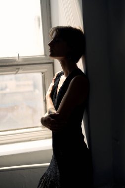 Siyah elbiseli zarif bir kadın. Kolları çaprazlanmış. Gün ışığında pencerenin önünde duruyor.