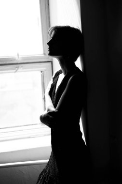 Siyah elbiseli bir kadın silueti. Parlak pencerenin yanında çapraz kollar arasında dramatik gölgeler yaratıyor.