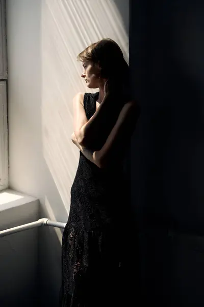 Siyah elbiseli kadın minimalist iç dekorda parlak pencerenin önünde duruyor.