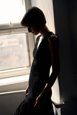 Siyah elbiseli kadın zarif bir şekilde pencere kenarında oturuyor. Yumuşak doğal ışıklandırmayla.