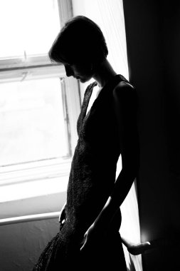 Siyah elbiseli kadının sanatsal silueti dramatik kontrastlı aydınlatmayla pencerede oturuyor