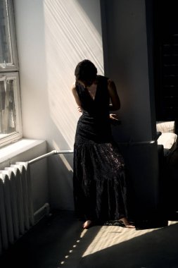 Siyah elbiseli kadın, dramatik gölgeleri ve doğal ışıkla pencerenin kenarında oturuyor.