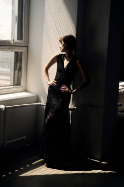 Siyah elbiseli kadın geniş pencerenin yanında oturuyor ve içeri ışık giriyor.