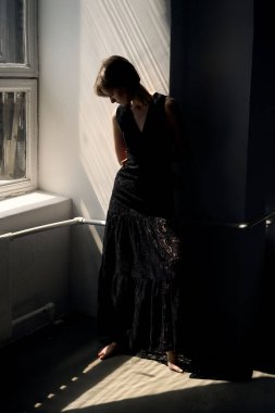 Siyah elbiseli bir kadın, parlak pencerenin kenarında, düşüncelere dalmış, gün ışığında oturuyor.