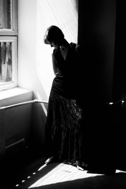 Siyah elbiseli, pencere arkası ışıklandırmalı dramatik siluet bir kadın portresi atmosferik atmosfer yaratıyor.
