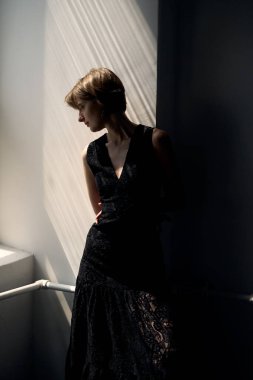Siyah kolsuz elbiseli kadın pencerenin kenarında narin doğal ışıklandırmayla duruyor.