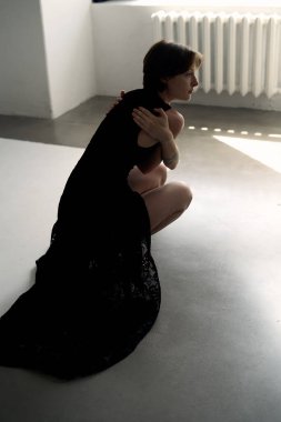 Geleneksel beyaz radyatörün yanında oturan siyah elbiseli bir kadının minimalist stüdyo fotoğrafı.