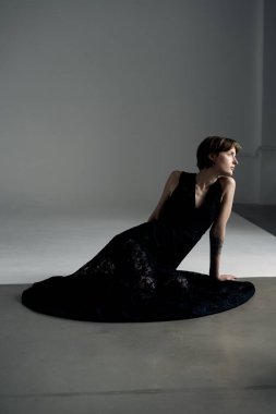 Akan siyah elbiseli zarif bir kadın aydınlanmış beyaz stüdyo zemininde uzanır.