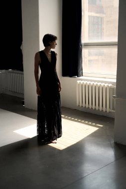 Dalgalı siyah elbiseli zarif bir kadın minimalist iç mekanda güneşli pencerenin yanında duruyor.