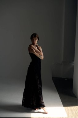 Minimalist stüdyo ortamında kollarını kavuşturmuş uzun siyah elbiseli zarif bir kadın.