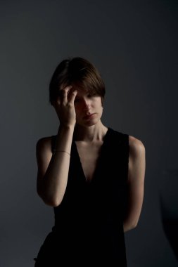 Siyah elbiseli, alnına dokunan genç esmer kadın. Tarafsız bir arkaplanda parmaklarıyla düşünceli bir poz vermiş.