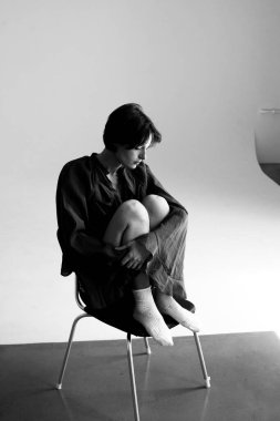 Minimalist stüdyo portresi, siyah ceketli, beyaz koltukta oturan, arka planı temiz.