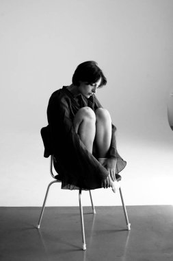 Huysuz stüdyo portresi, siyah ceketli, beyaz koltuğa yerleştirilmiş dramatik ışıklandırmalı.