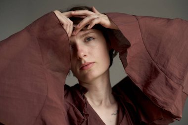 Pas rengi büyük tişörtlü zarif bir kadın. Elleri başının arkasında, stüdyo ışıkları altında poz veriyor.