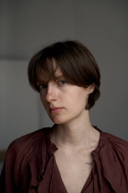 Kısa kahverengi saçlı, pas rengi bluzlu, kibar ifadeli ve hafif ışıklı bir kadının doğal portresi.