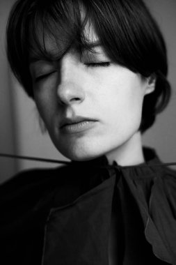 Kısa siyah saçlı, minimalist stüdyo ortamında zarif siyah bir bluz giyen genç bir kadının dramatik siyah beyaz portresi.