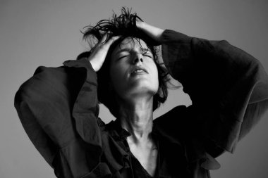 Kıvırcık saçlı ve dramatik stüdyo ışıklandırmasında kollarını kaldırmış bir kadının sanatsal siyah beyaz portresi.