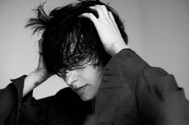 Tek renkli stüdyo portresi. Koyu renk kazaklı, elleri saçı dalgın bir şekilde duran genç bir adam.