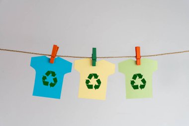 Açık sarı, mavi ve açık yeşil tişört şeklinde bir kağıt geri dönüşüm sembolü içeriyor, ipe asılı, sürdürülebilir moda ve çevre bilincini sembolize ediyor