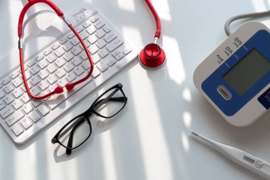 Doktorun sabahki masa konsepti; Kırmızı bir steteskop, dijital gerilim ve termometre üst görünümü, cam pencerelerden sabah ışığı altında ve klavye altında