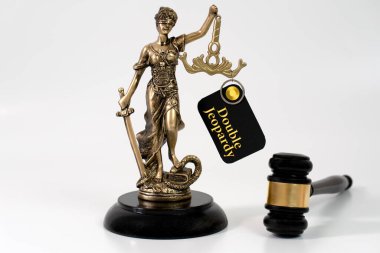 Bir Adalet Heykeli, 'Çifte Riziko' başlıklı bir kağıt ile bir tokmağın yanında duruyor. Aynı suçtan dolayı birden fazla kovuşturmaya karşı hukuki korumayı simgeliyor.