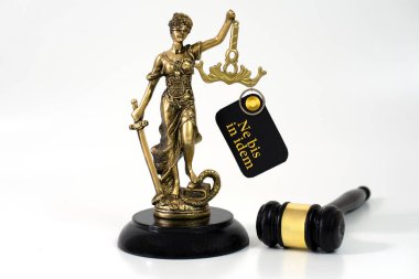 Bir Adalet Heykeli, elinde 'Ne Bis In İdem' başlıklı bir kağıt bulunan bir tokmağın yanında duruyor. Aynı suçtan birden fazla kovuşturmayı yasaklayan hukuki ilkeyi sembolize ediyor.