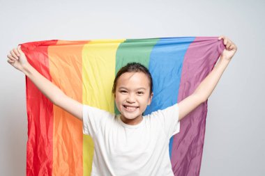 Genç bir kız gökkuşağı bayrağı tutuyor ve gülümsüyor. Kızın kimliğinde mutluluk ve gurur kavramı