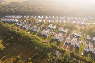 Yeşil bir ormanın yanında aynı beyaz evlere sahip modern bir konut arazisinin insansız hava aracı görüntüsü. Yerleşim yerleri, güneşin doğuşunun ya da batışının sıcacık ve puslu ışıklarıyla yıkanarak uzun gölgeler yaratır. Bu resim bu tür kavramları temsil eder