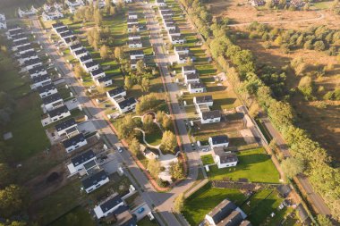 Yeşil bir ormanın yanında aynı beyaz evlere sahip modern bir konut arazisinin insansız hava aracı görüntüsü. Yerleşim yerleri, güneşin doğuşunun ya da batışının sıcacık ve puslu ışıklarıyla yıkanarak uzun gölgeler yaratır. Bu resim bu tür kavramları temsil eder