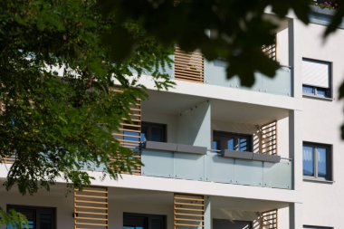 Güneşli bir günde modern bir apartmanın dışında. Ön cephede buzlu cam korkuluklar ve dekoratif ahşap çıtalarla çağdaş balkonlar yer alıyor. Önplandaki yeşil bir ağaç şehir ortamına doğanın bir dokunuşunu ekler. Modern için kavram