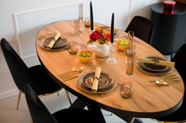 Modern bir yemek odasına zarif bir şekilde ahşap oval yemek masası koydu. Masa koyu tabaklar, altın çatal bıçak ve şık cam eşyalar ile bir akşam yemeği partisi için hazırlandı. Dekorda siyah mumlar, sonbahar buketi olan bir vazo ve fr ile kaseler yer alıyor.