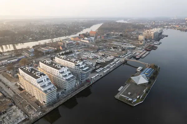 Güneşin doğuşunda ya da batışında şehrin insansız hava aracı görüntüsü. Şehir manzarasında modern konut binaları, birçok otobüsün park ettiği büyük bir otobüs durağı ve mavnalı bir liman alanı yer alıyor. Binası olan küçük bir ada da görülebilir. T