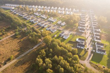 Yeşil bir ormanın yanında aynı beyaz evlere sahip modern bir konut arazisinin insansız hava aracı görüntüsü. Yerleşim yerleri, güneşin doğuşunun ya da batışının sıcacık ve puslu ışıklarıyla yıkanarak uzun gölgeler yaratır. Bu resim bu tür kavramları temsil eder