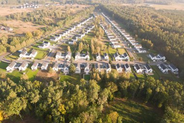 Yeşil bir ormanın yanında aynı beyaz evlere sahip modern bir konut arazisinin insansız hava aracı görüntüsü. Yerleşim yerleri, güneşin doğuşunun ya da batışının sıcacık ve puslu ışıklarıyla yıkanarak uzun gölgeler yaratır. Bu resim bu tür kavramları temsil eder