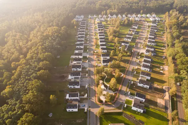 Yeşil bir ormanın yanında aynı beyaz evlere sahip modern bir konut arazisinin insansız hava aracı görüntüsü. Yerleşim yerleri, güneşin doğuşunun ya da batışının sıcacık ve puslu ışıklarıyla yıkanarak uzun gölgeler yaratır. Bu resim bu tür kavramları temsil eder