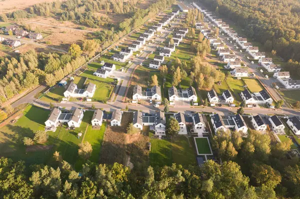 Yeşil bir ormanın yanında aynı beyaz evlere sahip modern bir konut arazisinin insansız hava aracı görüntüsü. Yerleşim yerleri, güneşin doğuşunun ya da batışının sıcacık ve puslu ışıklarıyla yıkanarak uzun gölgeler yaratır. Bu resim bu tür kavramları temsil eder