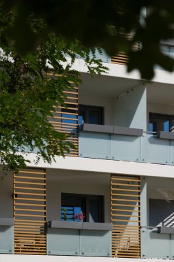 Güneşli bir günde modern bir apartmanın dışında. Ön cephede buzlu cam korkuluklar ve dekoratif ahşap çıtalarla çağdaş balkonlar yer alıyor. Önplandaki yeşil bir ağaç şehir ortamına doğanın bir dokunuşunu ekler. Modern için kavram